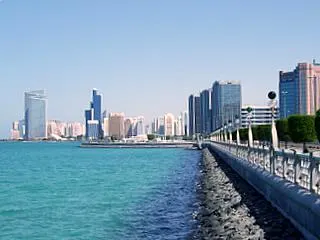 A orla da Corniche de Abu Dhabi, com a balaustrada branca à beira da água turquesa e a linha de prédios altos ao fundo.