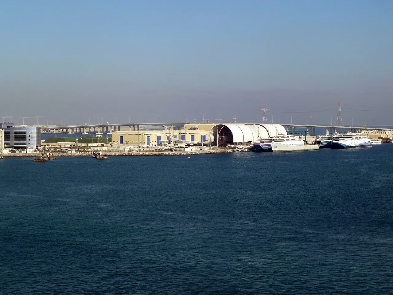 O porto de Mina Zayed visto da água, com galpões, guindastes e barcos atracados na margem, sob céu claro.