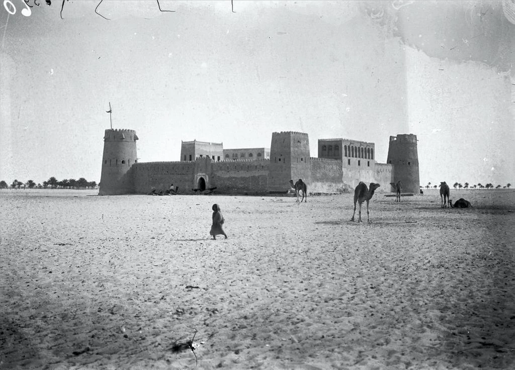 Fotografia histórica de 1904 de Qasr Al Hosn, em Abu Dhabi: o forte cercado de areia, com torres e camelos à frente.