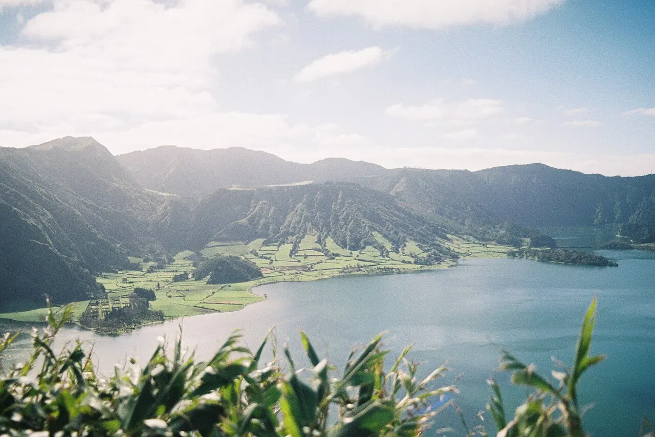 A Lagoa das Sete Cidades dentro da caldeira, com campos verdes na margem, as encostas da cratera e folhagem em primeiro plano.