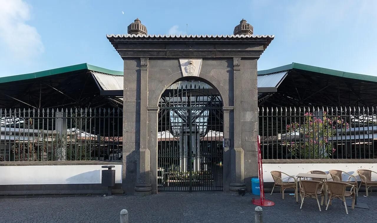 O portão de pedra escura do Mercado da Graça, com grade de ferro, a cobertura do mercado atrás e mesas de café na calçada.