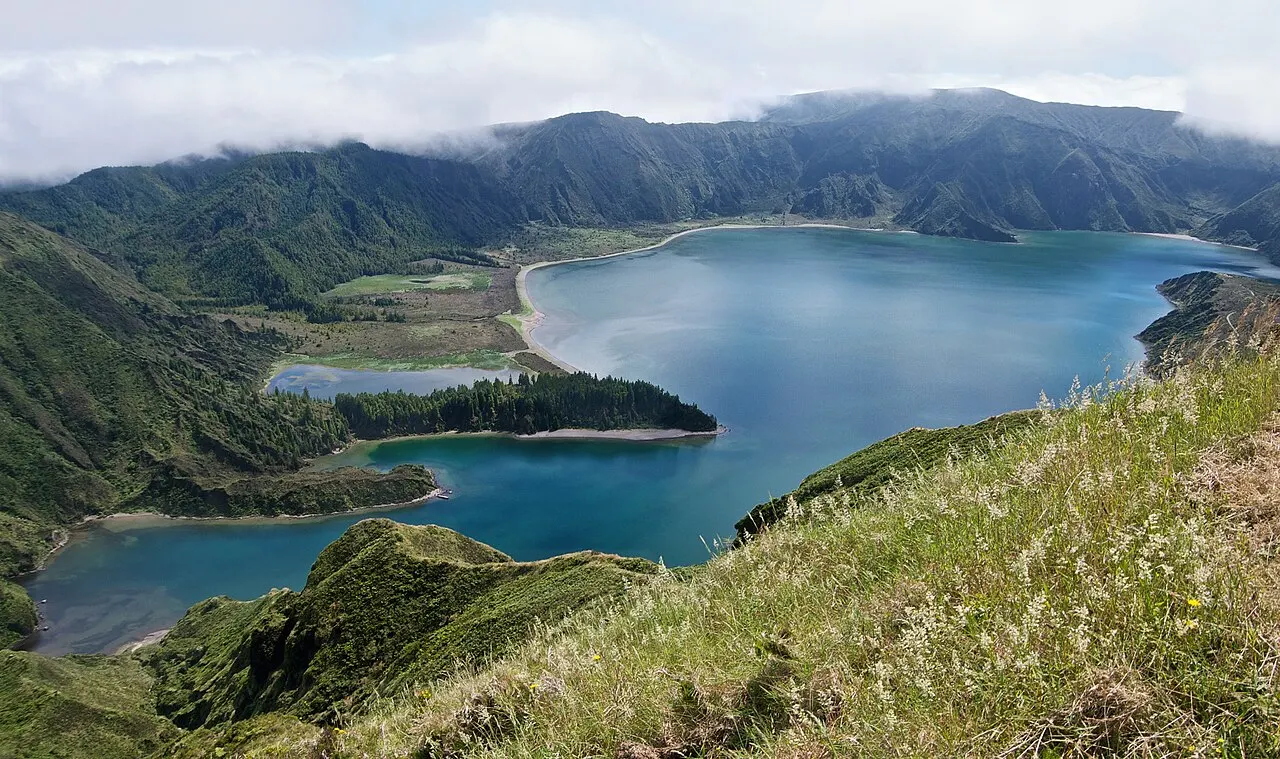 A Lagoa do Fogo azul dentro da cratera, com praias claras na margem e as encostas verdes com nuvens no alto.