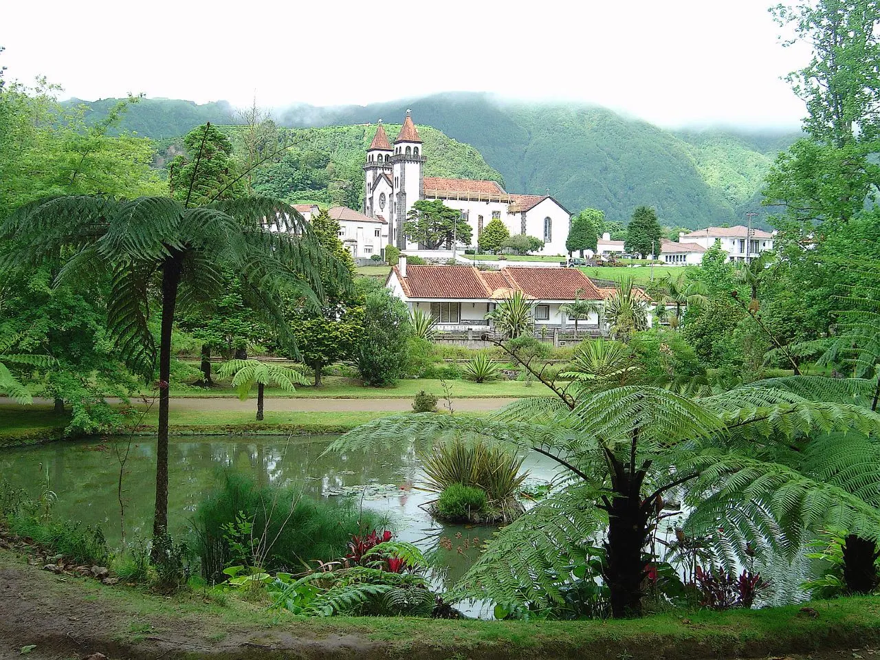 Um lago do Parque Terra Nostra entre fetos arborescentes, com as casas brancas e a igreja das Furnas ao fundo.