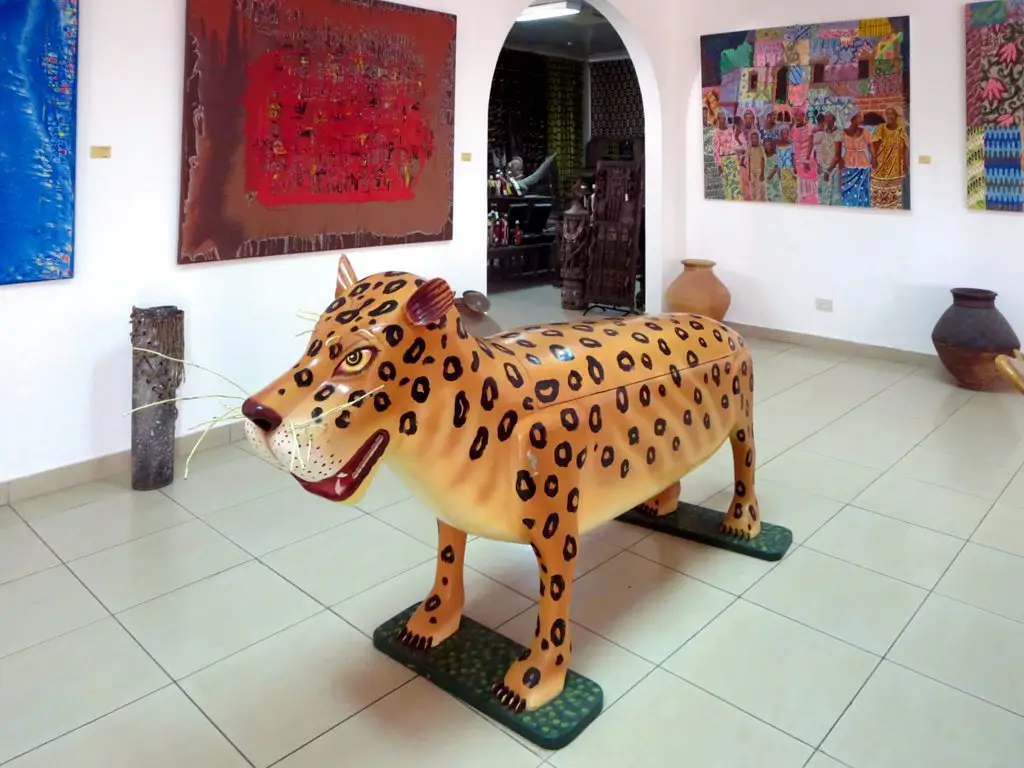 Caixão de madeira pintado como um felino com manchas, no meio de uma sala branca com quadros.