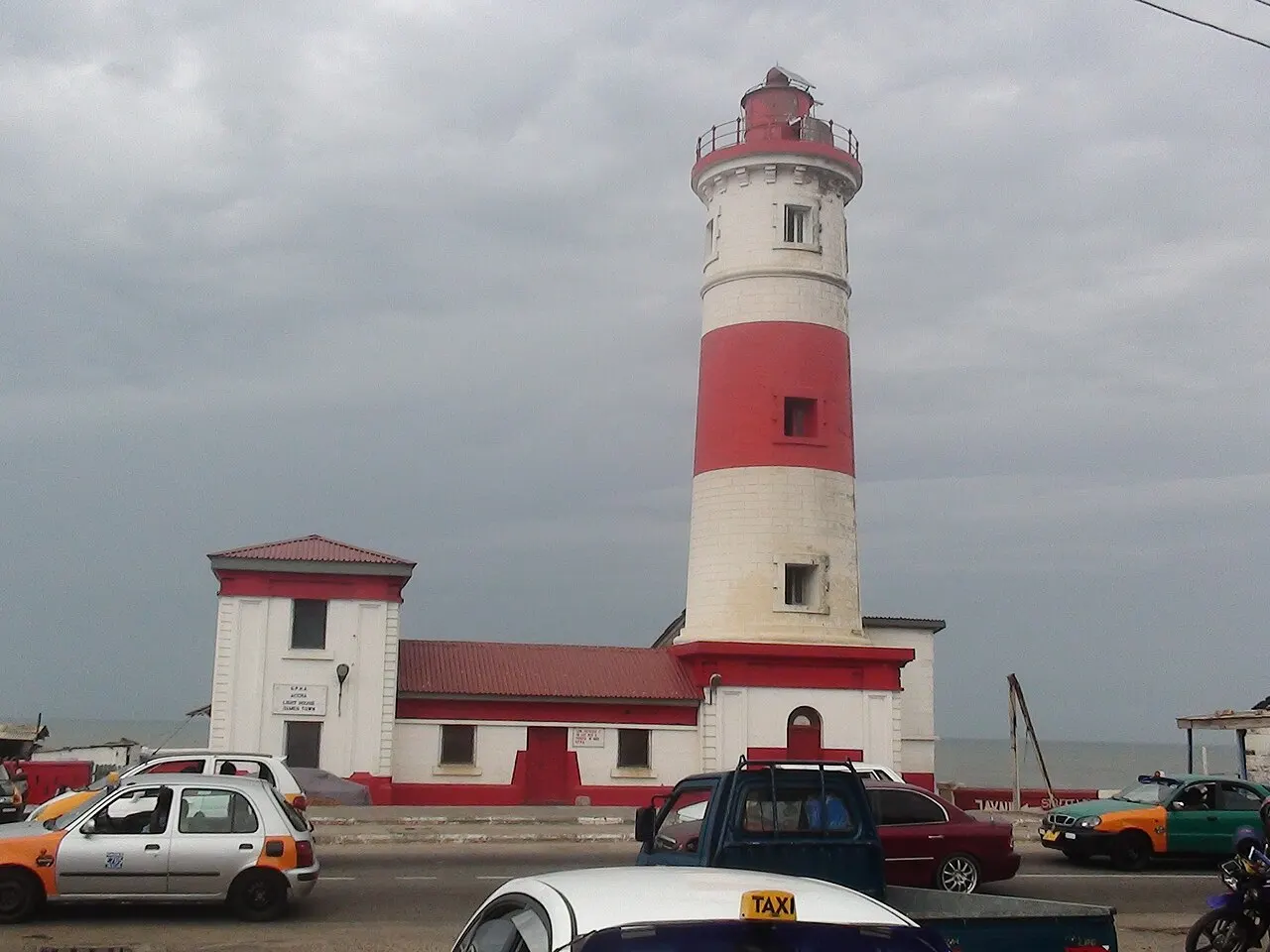 Farol de Jamestown, em Acra, listrado de vermelho e branco, acima de casas baixas e carros.