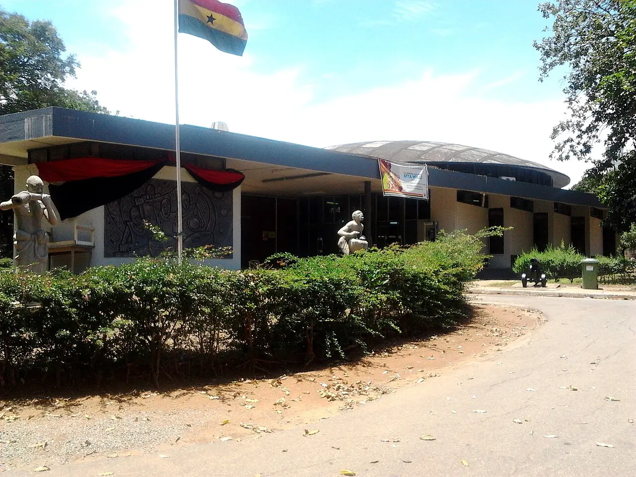 Edifício baixo do museu com cobertura curva, esculturas junto à entrada, uma bandeira de Gana e vegetação em primeiro plano.
