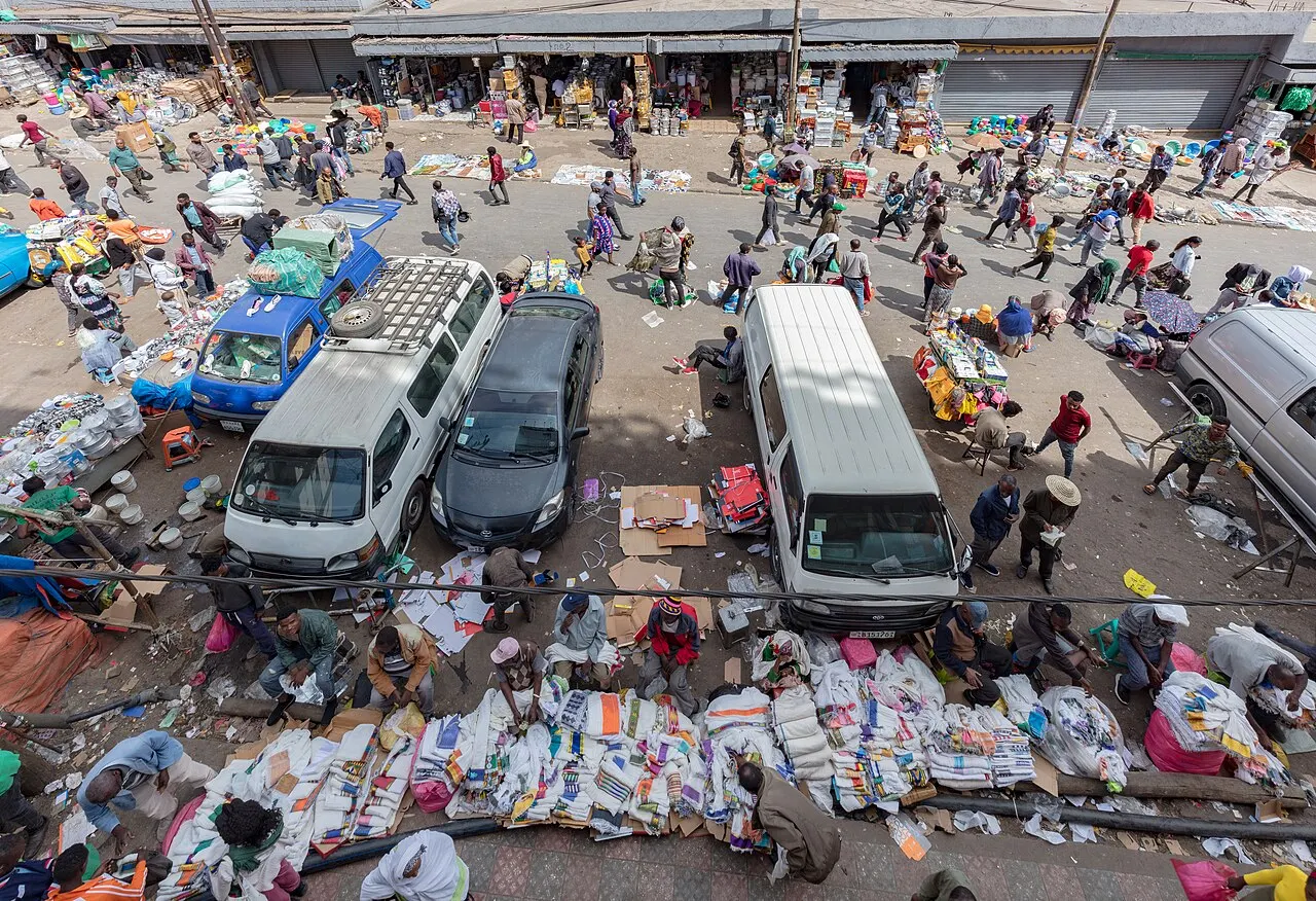 Vista de cima de uma rua de mercado, com furgões e um carro estacionados no meio, vendedores sentados junto a pilhas de tecidos dobrados no chão e pedestres circulando entre as barracas, em pleno dia.