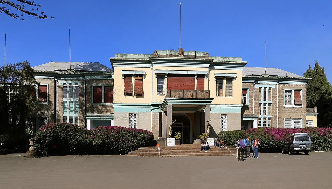 A fachada de um prédio histórico de dois andares, em amarelo e cinza, com uma escadaria central, jardins floridos e algumas pessoas paradas na entrada, sob céu azul de dia.