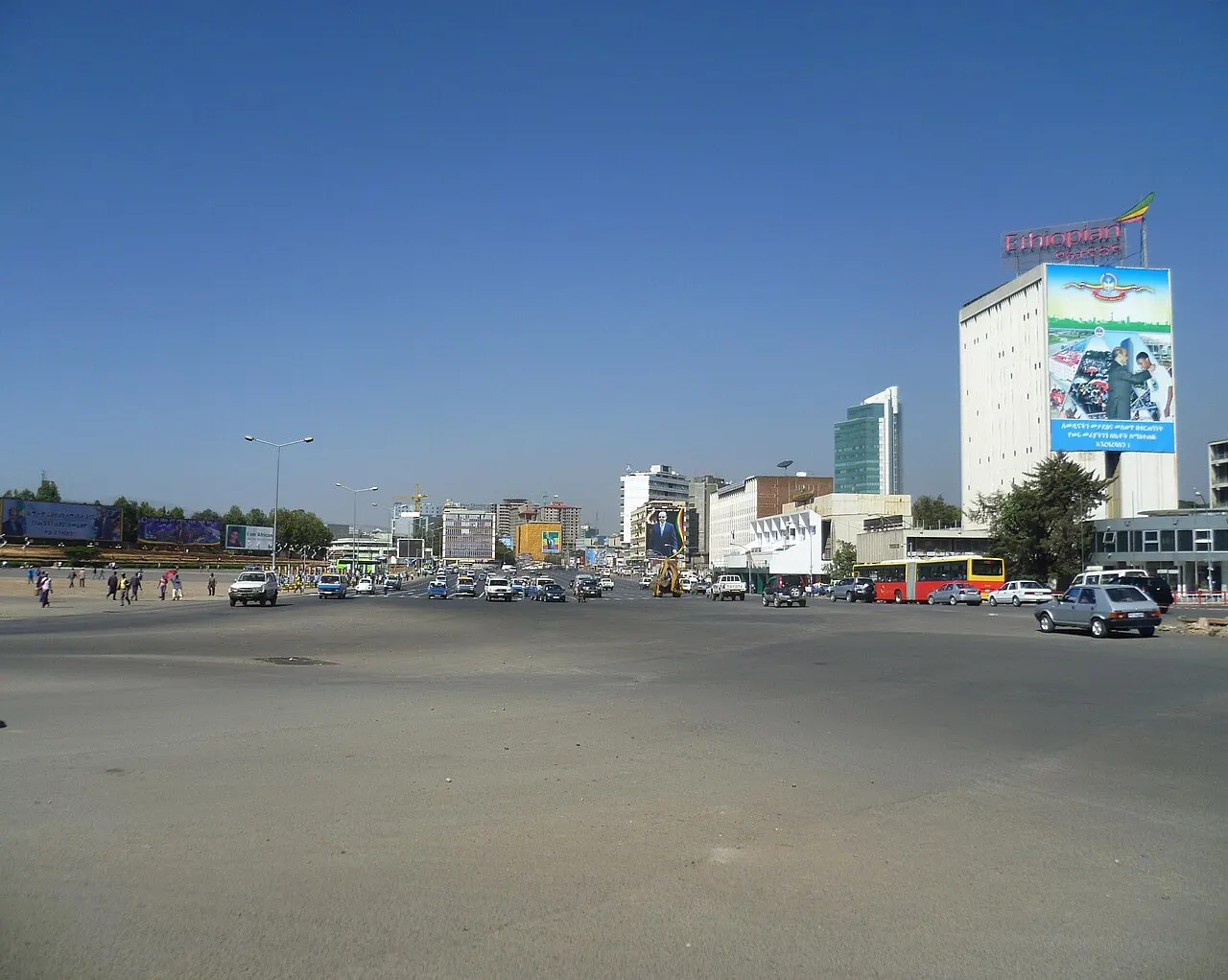 Largo asfaltado da Praça Meskel, com carros espalhados e um edifício branco de cartaz grande à direita, sob céu azul.