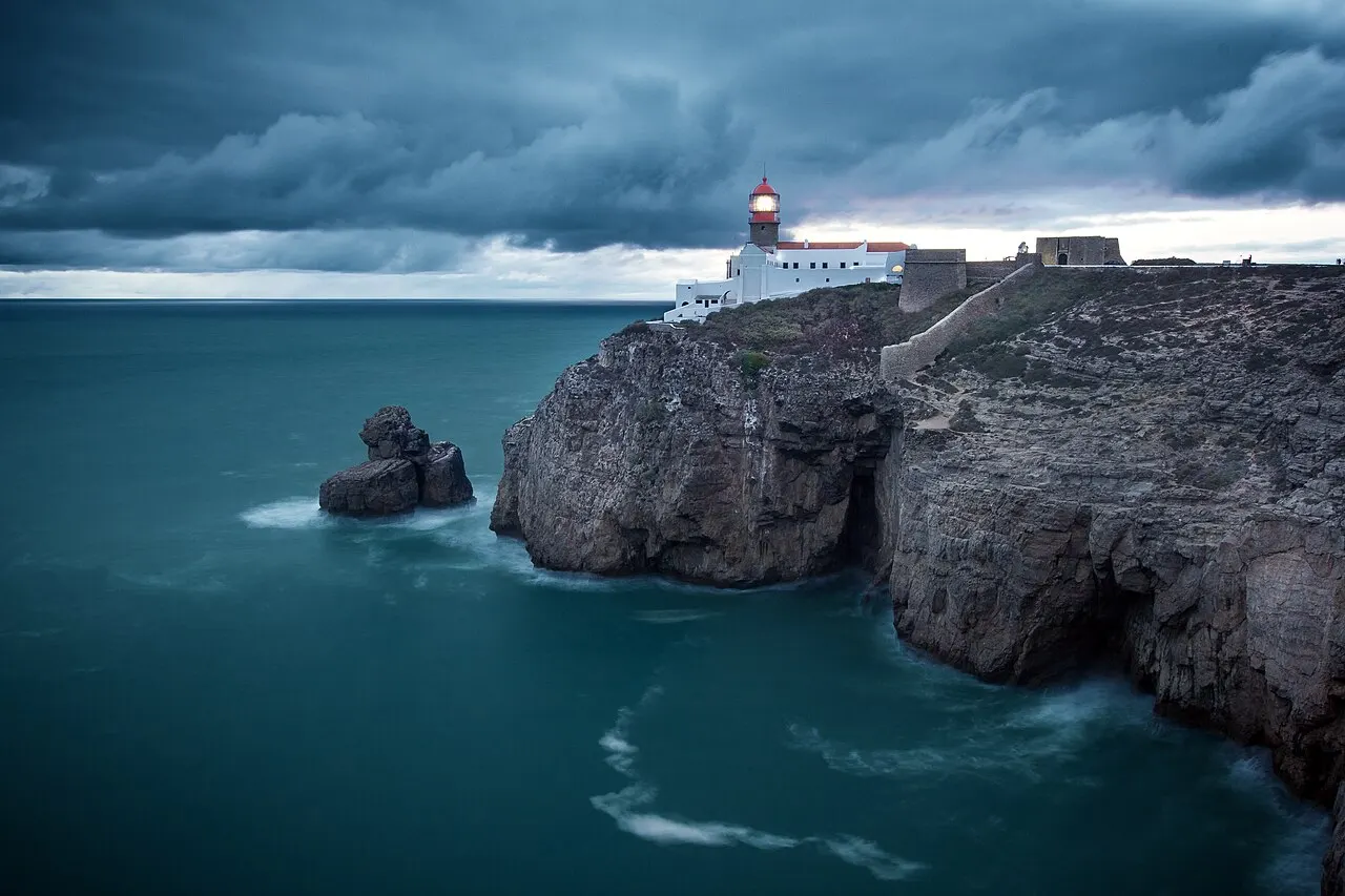 O farol do Cabo de São Vicente no alto de uma falésia escura sobre o mar verde-escuro, sob nuvens pesadas.