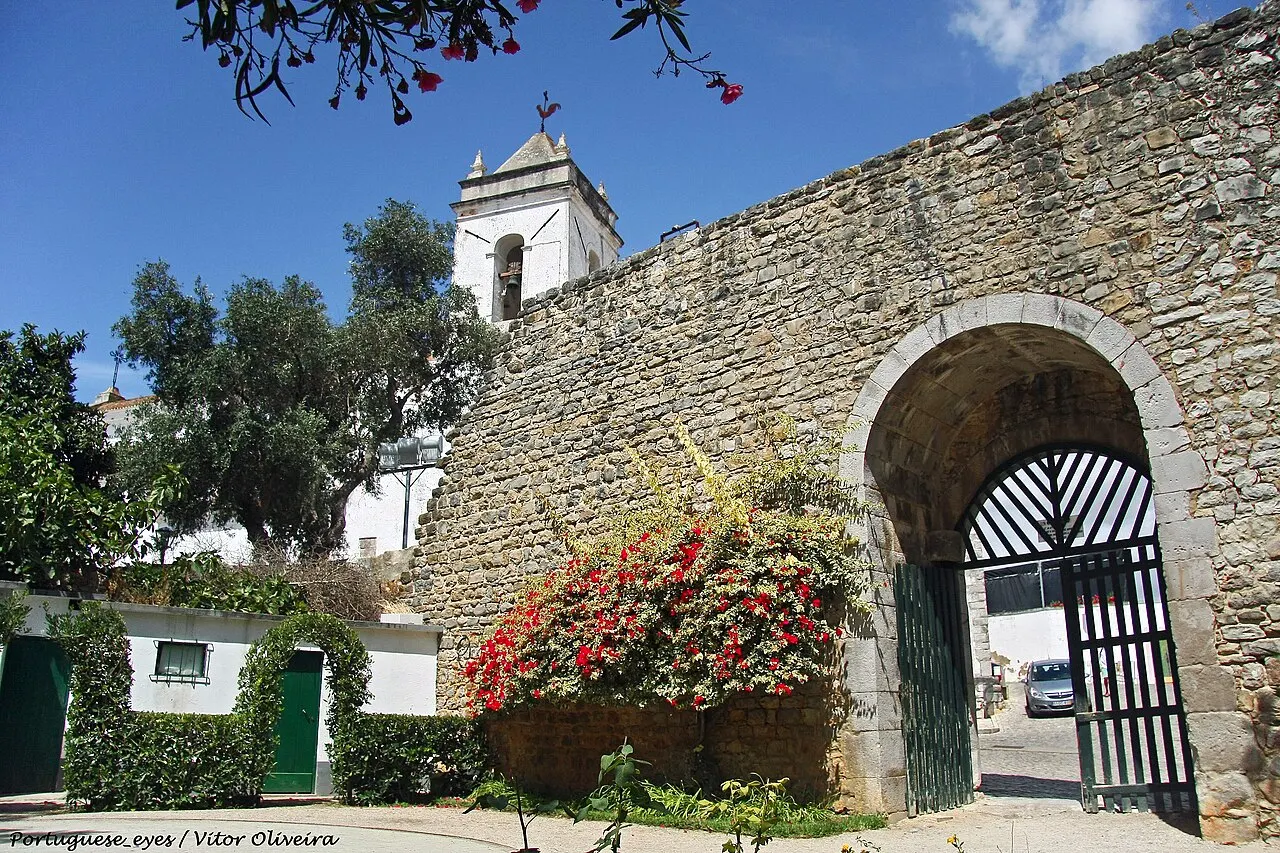 A muralha de pedra do Castelo de Tavira com uma porta em arco, buganvílias vermelhas e a torre de uma igreja atrás.