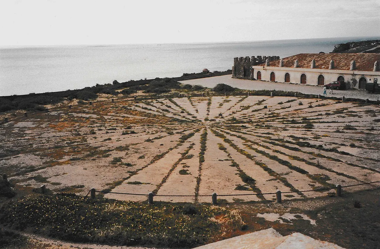 O grande círculo de pedras da rosa-dos-ventos dentro da Fortaleza de Sagres, com a muralha e o mar ao fundo.