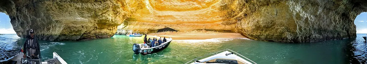 Panorâmica do interior da Gruta de Benagil: a abóbada de rocha dourada, a pequena praia lá dentro e barcos de passeio na água verde.