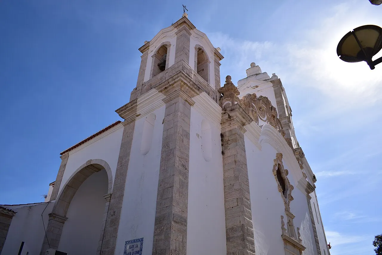 A fachada branca da Igreja de Santo António, em Lagos, com cantaria de pedra clara e a torre do sino, vista de baixo contra o céu azul.