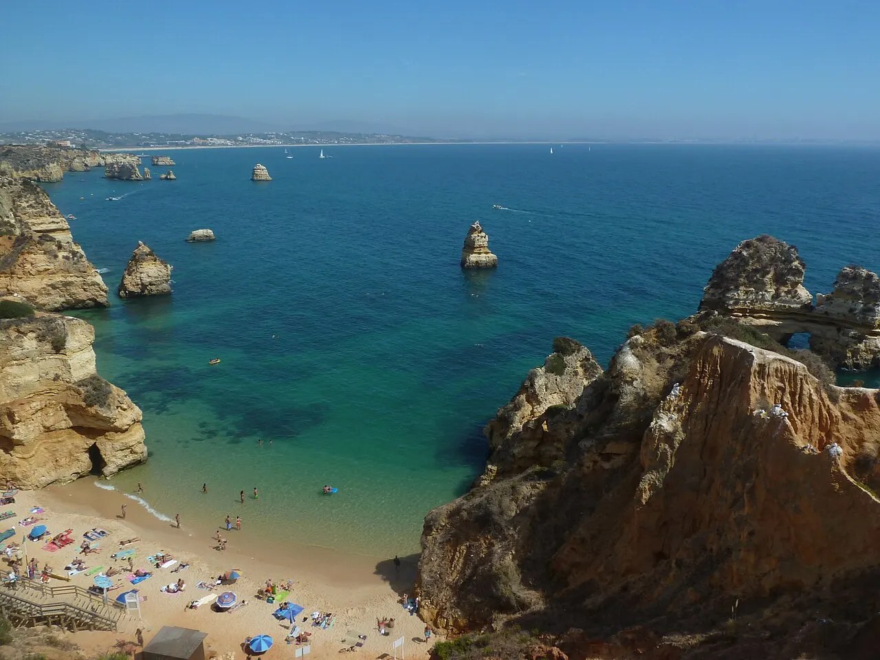A Praia do Camilo vista do alto, uma faixa pequena de areia cheia de guarda-sóis entre falésias douradas e o mar turquesa.