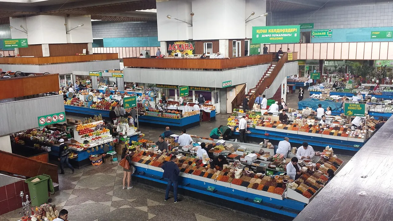 O interior de um mercado coberto de dois níveis, com bancas de frutas secas, nozes e frutas frescas dispostas em fileiras coloridas, escada de acesso ao mezanino e algumas pessoas circulando sob luz artificial.