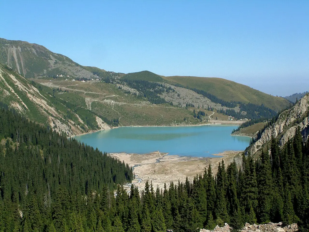 Lago turquesa do Big Almaty Lake no vale, entre pinheiros escuros e morros, sob céu azul.