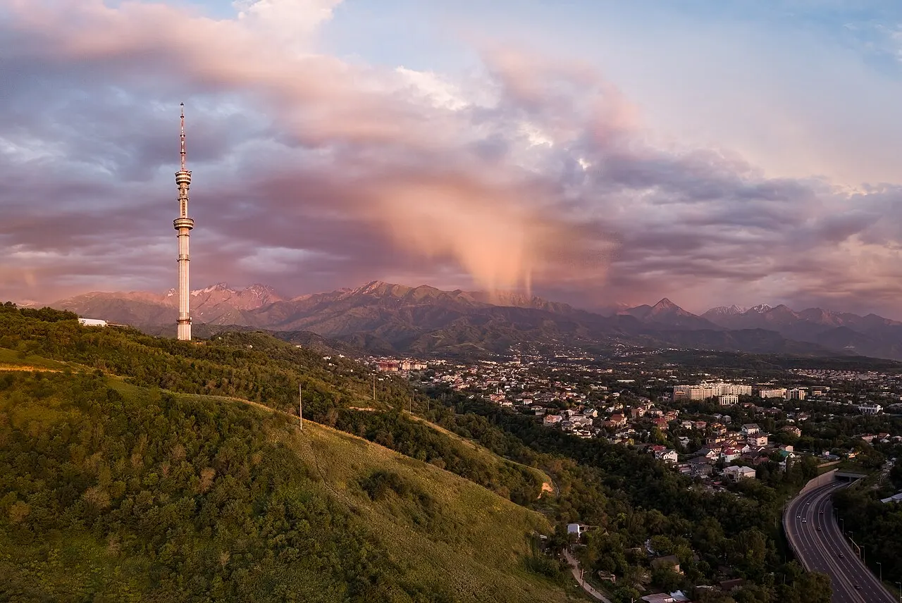 A torre de televisão do Kok-Tobe numa colina verde sobre Almaty, com a cidade baixa à direita e a cordilheira nevada do Alatau sob nuvens rosadas do fim do dia.