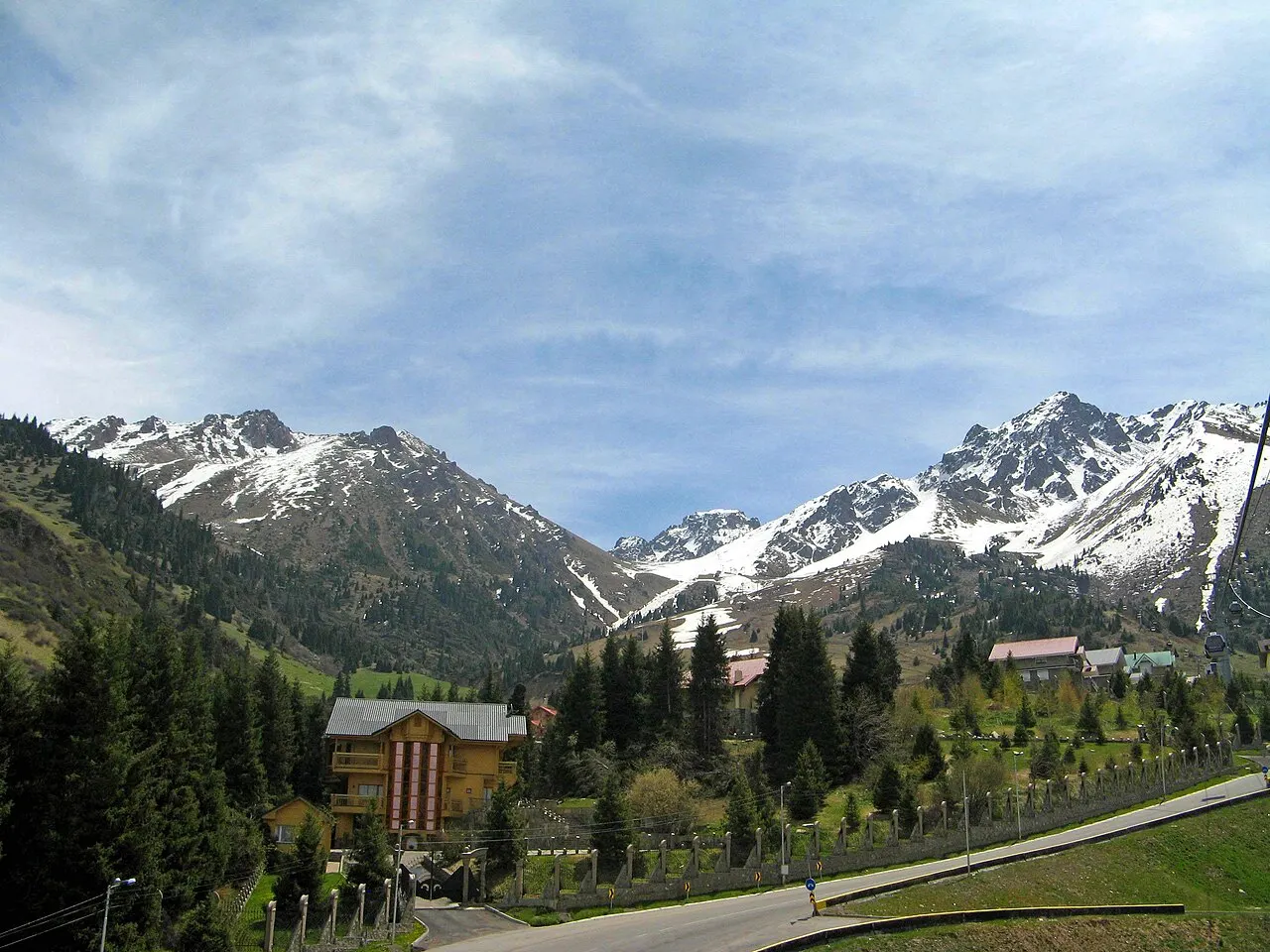 Um vale de montanha com picos nevados ao fundo, encostas cobertas de pinheiros, chalés de arquitetura alpina espalhados entre as árvores e uma estrada pavimentada em primeiro plano, sob céu claro.