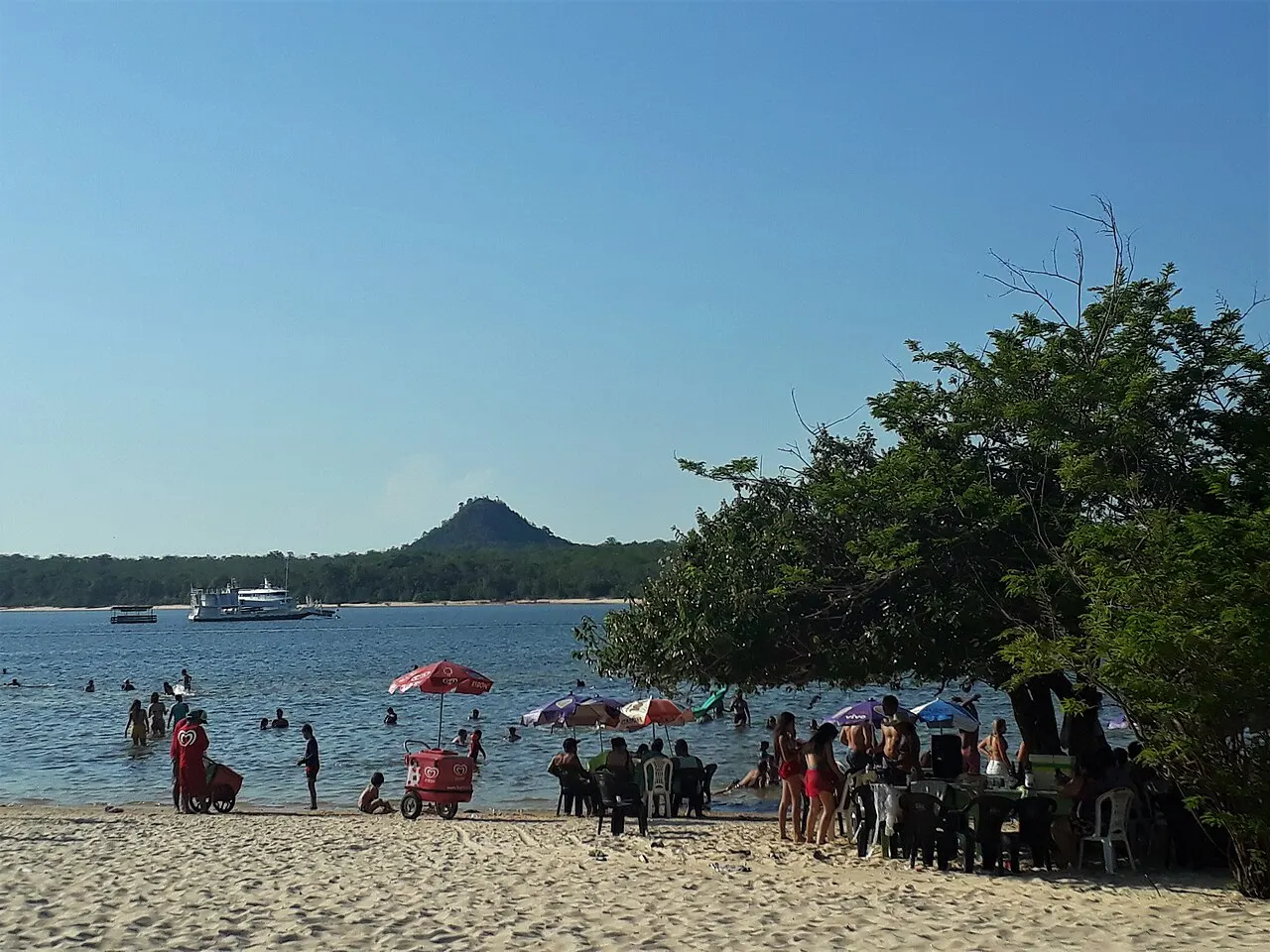 A praia da vila com banhistas e guarda-sóis, a água do Tapajós e, do outro lado, o morro pontudo da Serra da Piraoca.
