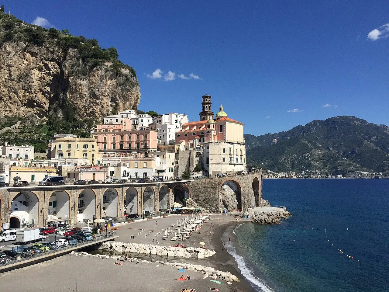 Atrani, com casas brancas e coloridas encostadas na falésia, uma igreja com campanário, os arcos da estrada costeira e a pequena praia de areia escura.