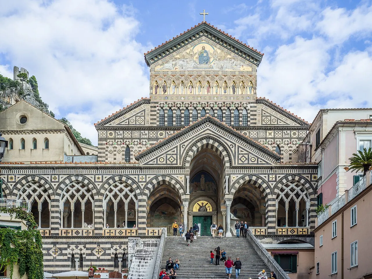A fachada do Duomo de Amalfi, com arcos e listras brancas e escuras, mosaico dourado no frontão e a escadaria larga cheia de visitantes.