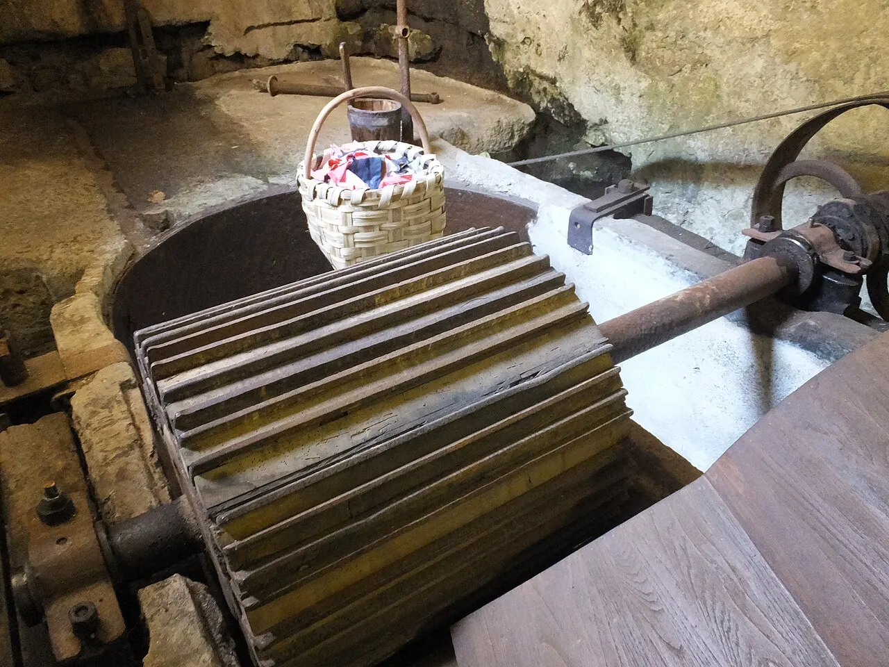Um cilindro de madeira com lâminas e engrenagens de ferro do antigo moinho de papel, dentro do Museo della Carta, em Amalfi, com paredes de rocha ao fundo.