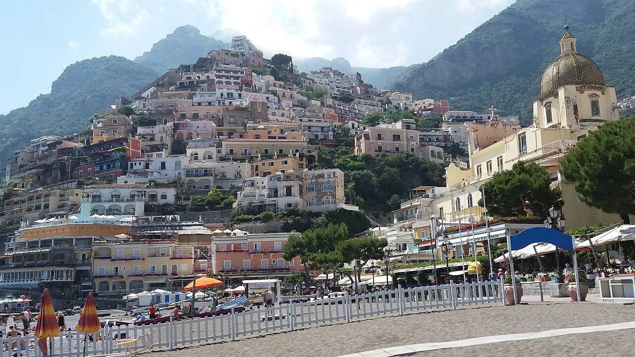 Positano vista da Spiaggia Grande, com o calçadão e guarda-sóis em primeiro plano e as casas coloridas subindo a encosta até a montanha.