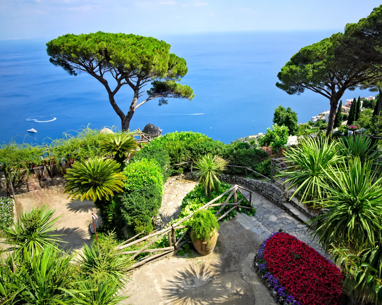 Os jardins da Villa Rufolo, com caminhos e cercas de madeira, canteiros floridos e pinheiros diante do mar azul.