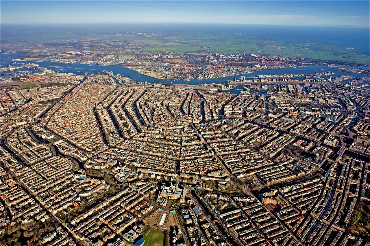 Vista aérea de uma cidade cortada por canais dispostos em curvas concêntricas, com prédios densos entre as vias de água e um trecho de mar e campos verdes no horizonte, em dia claro.