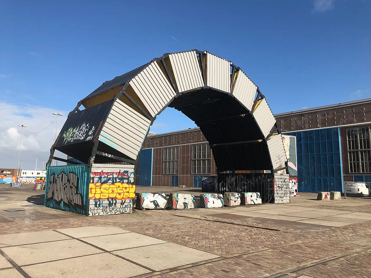 Arco de metal pintado no antigo estaleiro NDSM, em Amsterdã, coberto de cartazes e grafite, sob céu azul.