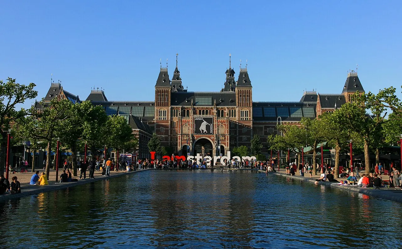 A fachada de tijolos vermelhos do Rijksmuseum, com torreões e telhados escuros, vista do outro lado de um espelho d'água onde pessoas sentam nas bordas, ao entardecer.