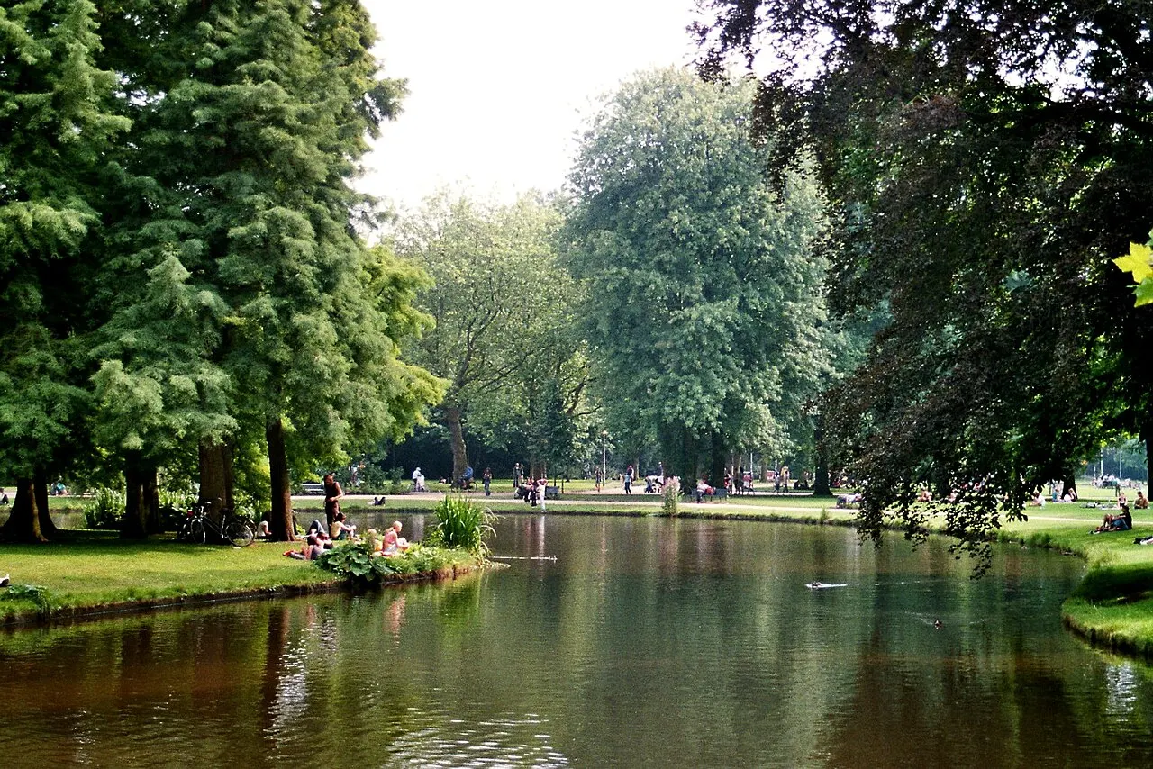 Lago do Vondelpark entre árvores altas, com pessoas sentadas na margem.