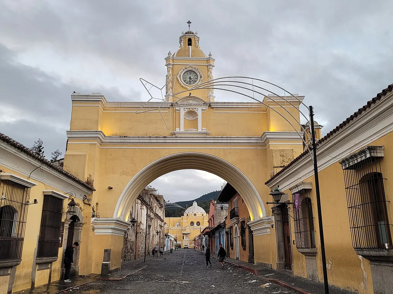 Um arco amarelo com uma torre de relógio no topo, atravessando uma rua de paralelepípedos ladeada por casas baixas coloniais, com uma igreja amarela ao fundo, sob céu nublado.