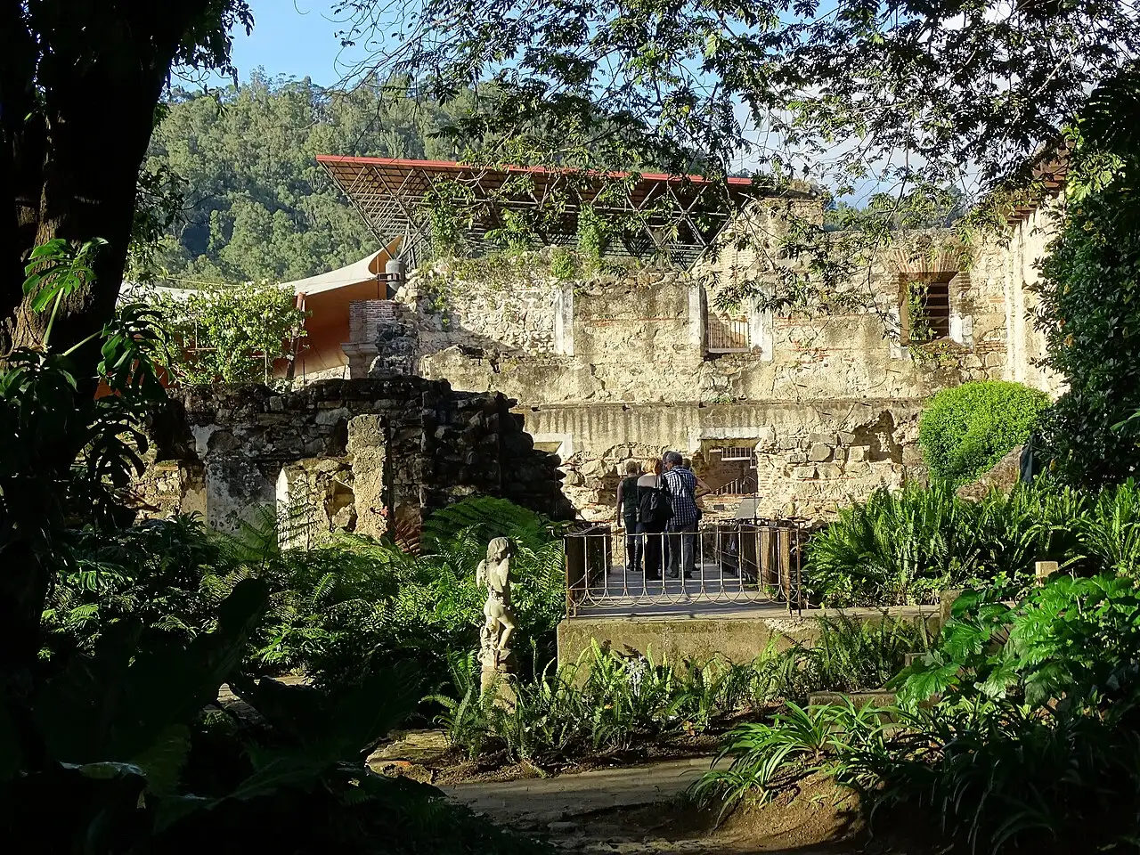Ruínas de pedra da Casa Santo Domingo no jardim, com um trecho de telhado novo e o morro ao fundo.