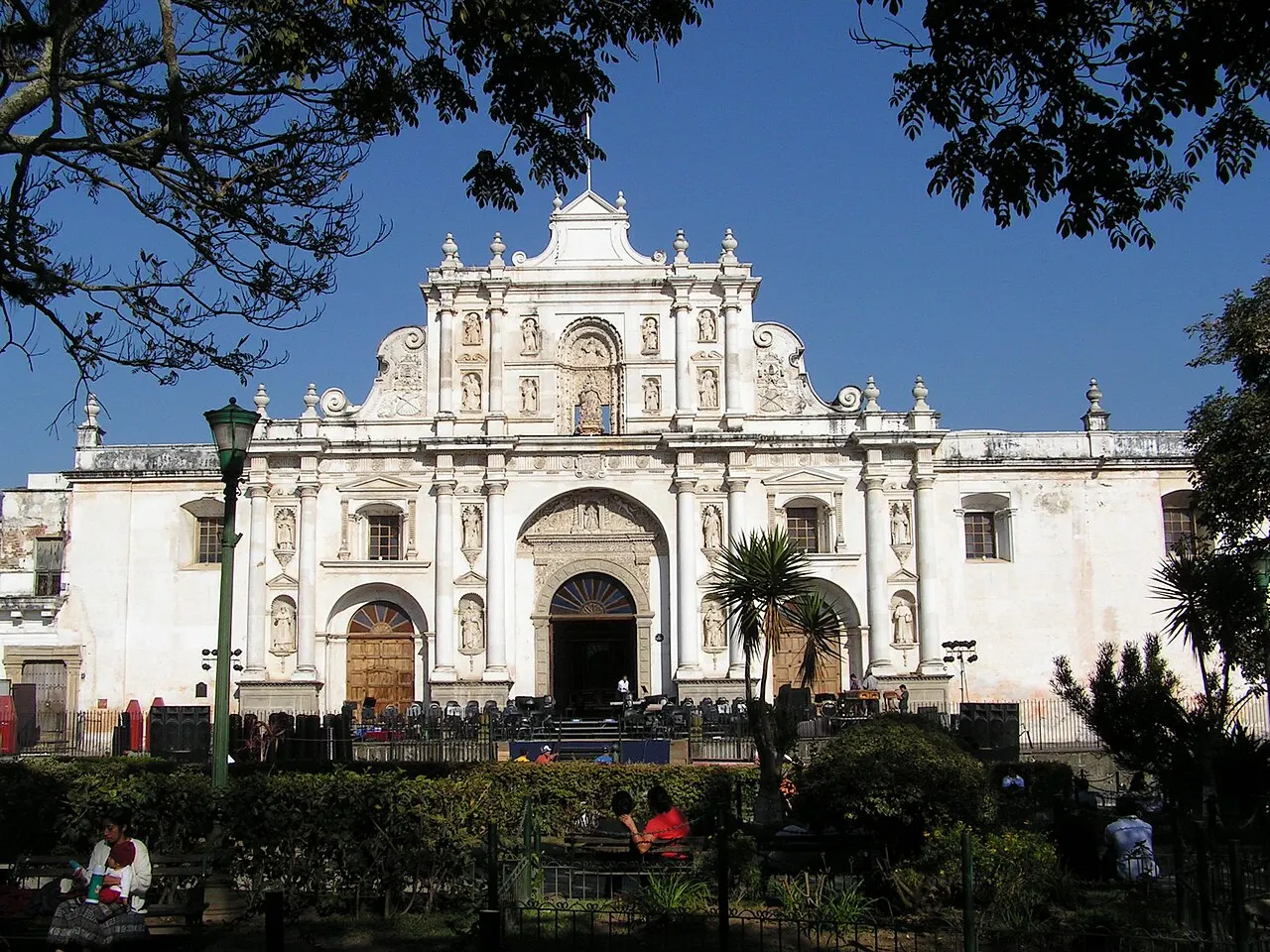 Fachada branca de estuque da catedral de San José, em Antígua, com nichos de santos, colunas e volutas, vista do parque central entre árvores e palmeiras.