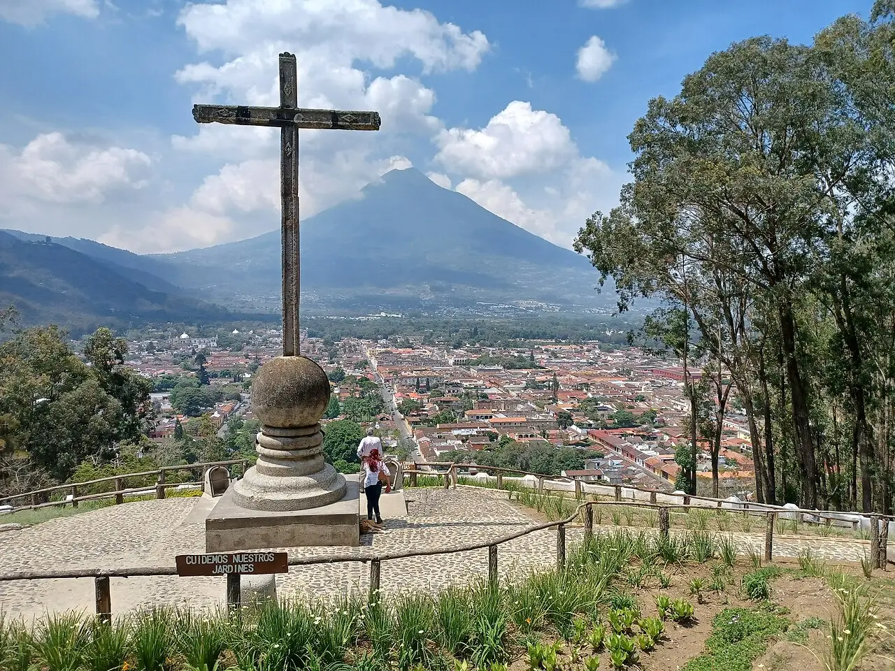 Uma cruz de pedra sobre um pedestal redondo, com os telhados de Antigua e um vulcão por trás das árvores.