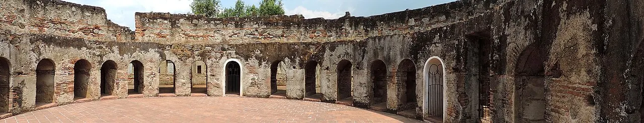Pátio circular de pedra do Convento de las Capuchinas, cercado por celas em ruínas com portas em arco e vegetação ao fundo sob céu claro.