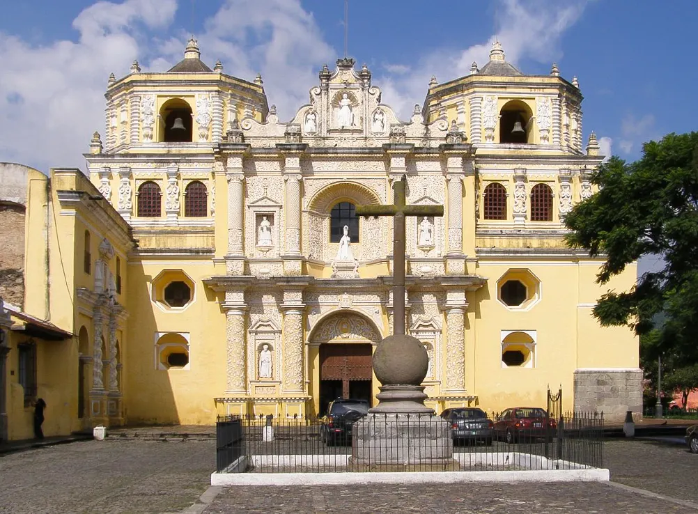 Igreja amarela de duas torres, com a cruz de pedra no adro empedrado.