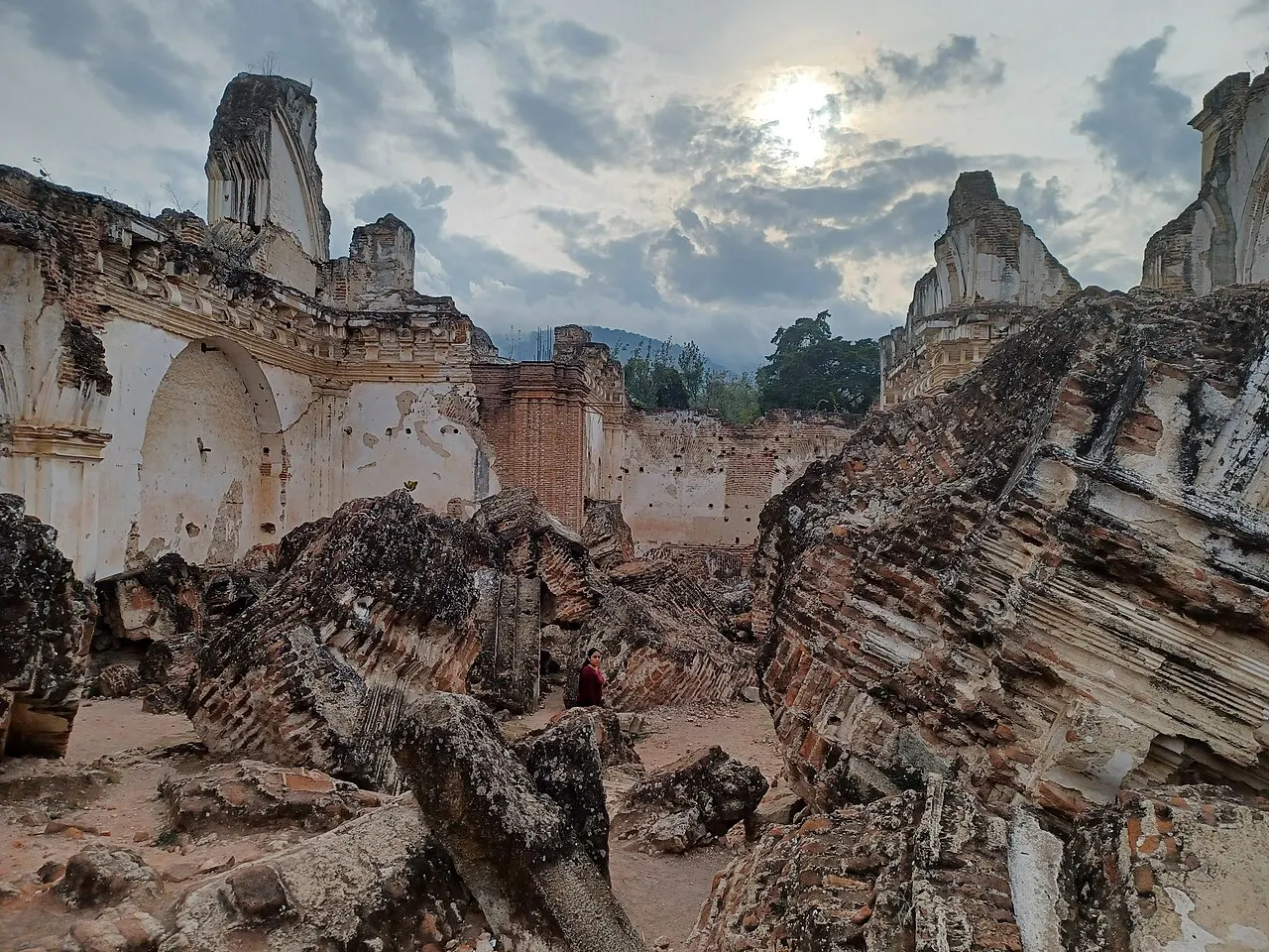 Interior sem teto do convento da Recolección, em Antígua: blocos inteiros da abóbada de tijolo caídos de lado no chão da nave, paredes altas de estuque branco rachado em volta e a serra atrás, sob céu nublado.