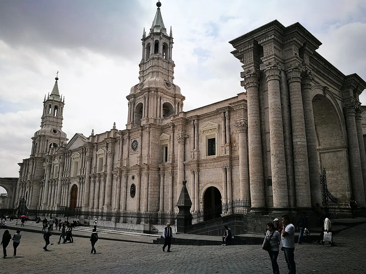 Fachada clara e extensa com colunas e duas torres, vista em diagonal a partir da praça, com pedestres no calçamento em primeiro plano.