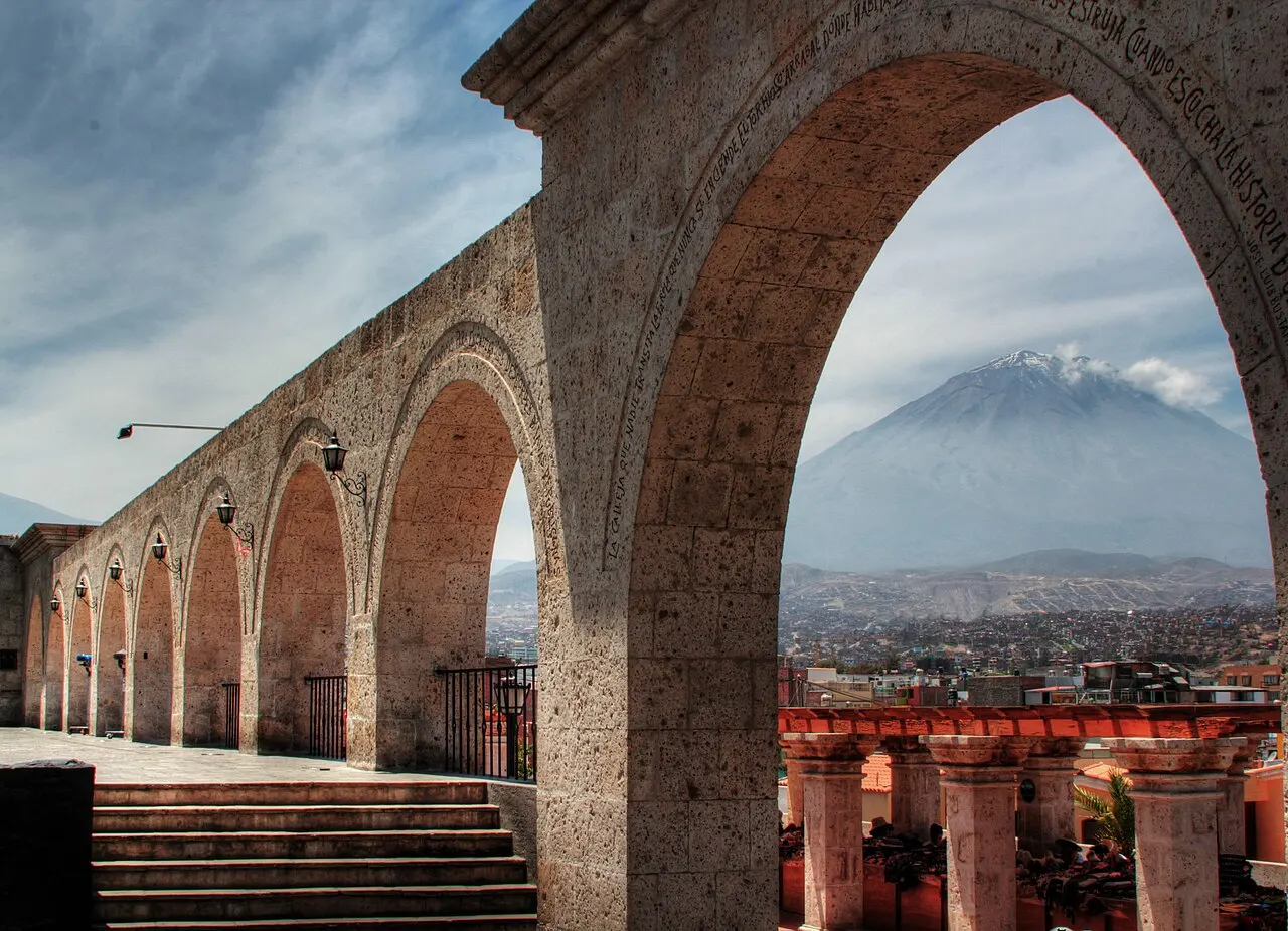 Galeria de arcos de pedra com inscrições; pelo arco maior aparece um vulcão de pico nevado acima da cidade.