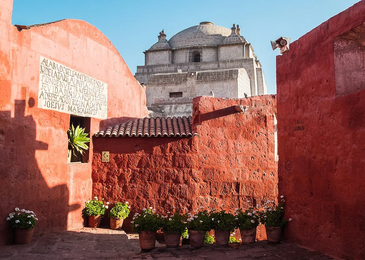 Muros pintados de vermelho, vasos de flores junto à parede, uma placa com inscrição e uma cúpula de pedra clara ao fundo, sob céu azul.