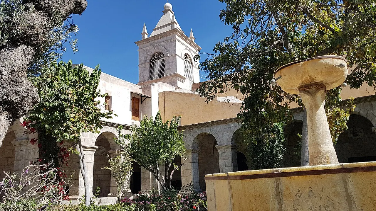 Claustro com arcos, árvores e flores, uma fonte de pedra em primeiro plano à direita e um campanário branco ao fundo.