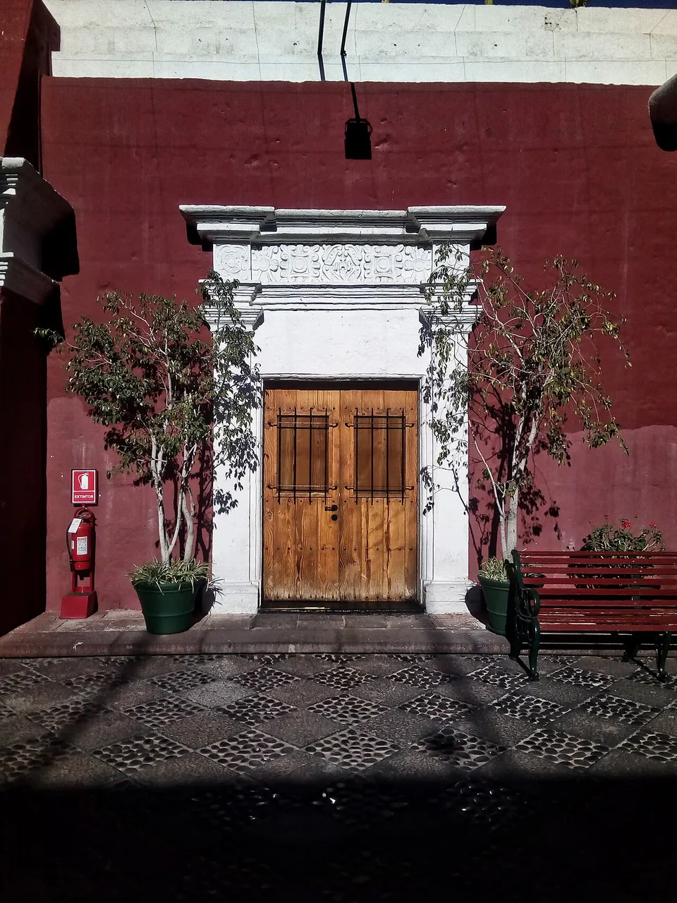 Porta de madeira com moldura de pedra branca lavrada, num muro vermelho, entre duas árvores em vasos, com um banco à direita e piso de pedras em desenho.