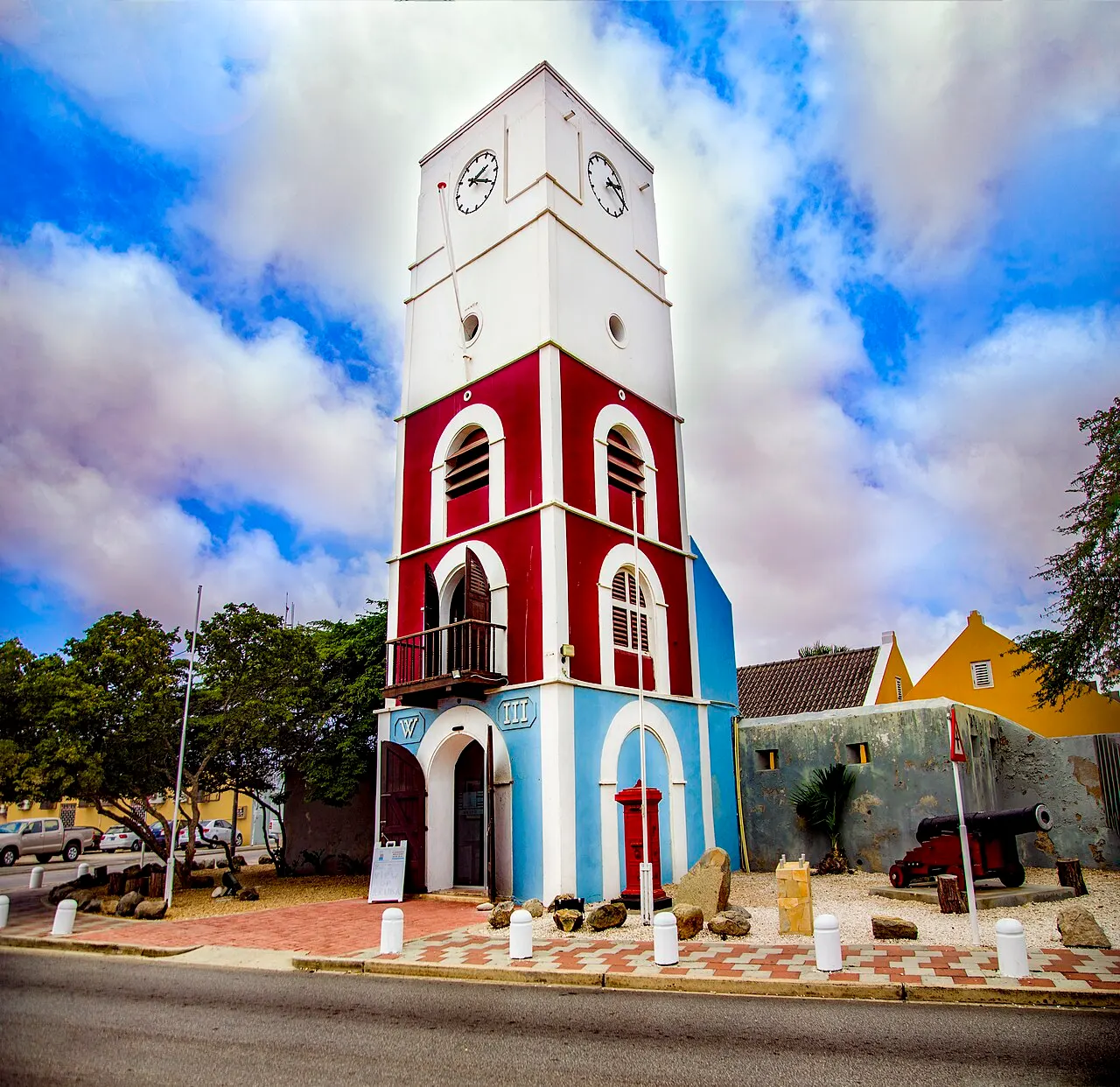 A torre Willem III do Fort Zoutman, em Oranjestad, pintada de branco, vermelho e azul, com um relógio no alto e céu com nuvens.
