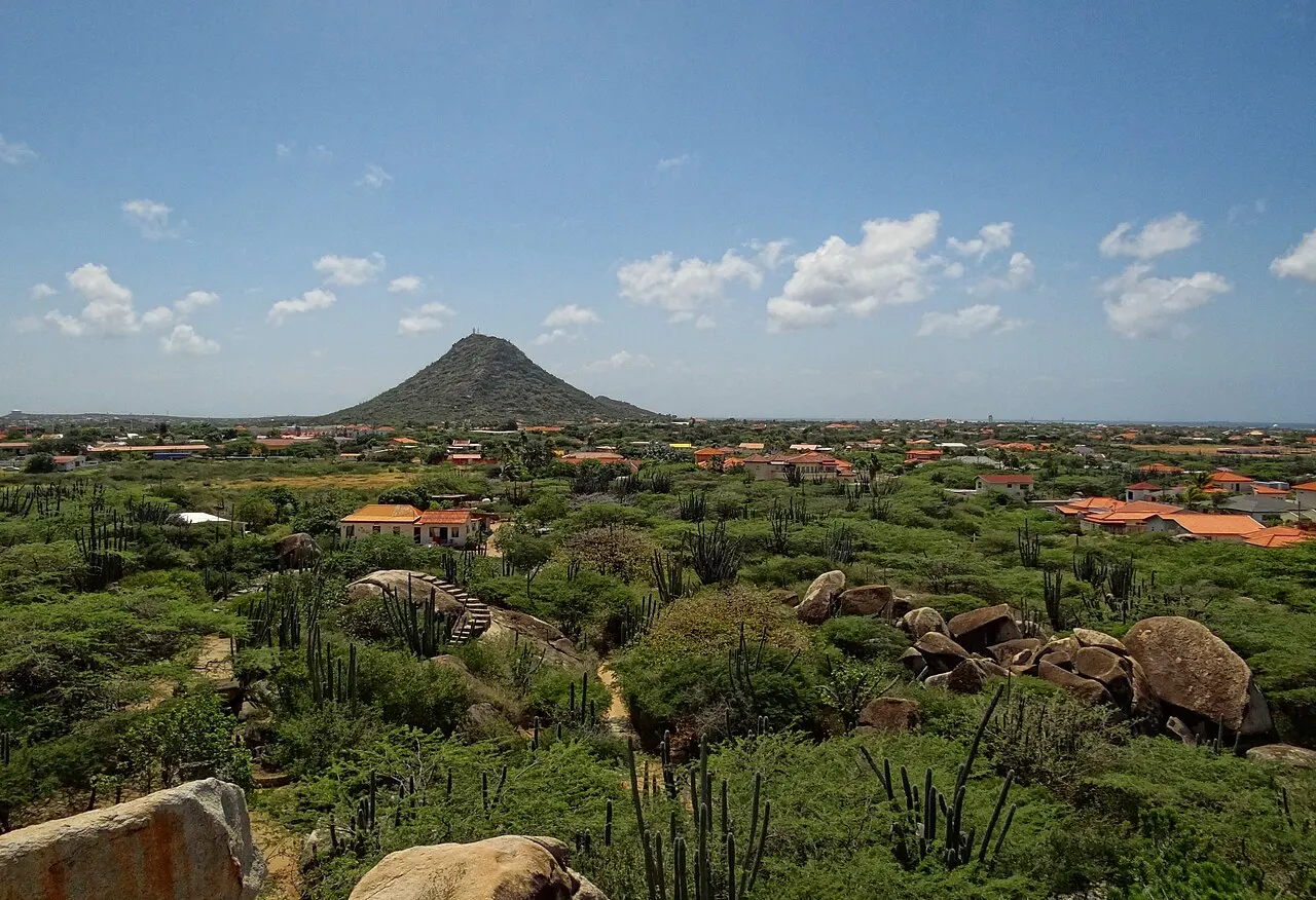 El cerro en forma de cono del Hooiberg en el horizonte, sobre una llanura de matorral y casas dispersas, bajo un cielo con nubes.