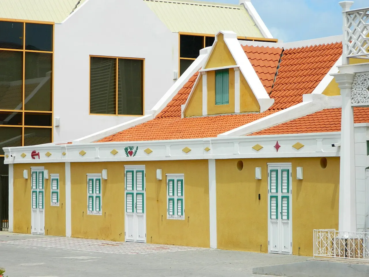 La fachada del Museo Arqueológico Nacional, en Oranjestad: una casa antigua amarilla con puertas verdes y techo naranja, junto a un edificio de vidrio.