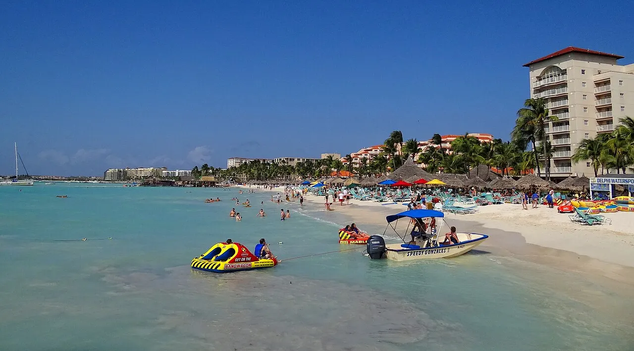 A água rasa e turquesa de Palm Beach com boias e barcos, guarda-sóis na areia e um hotel alto à direita.
