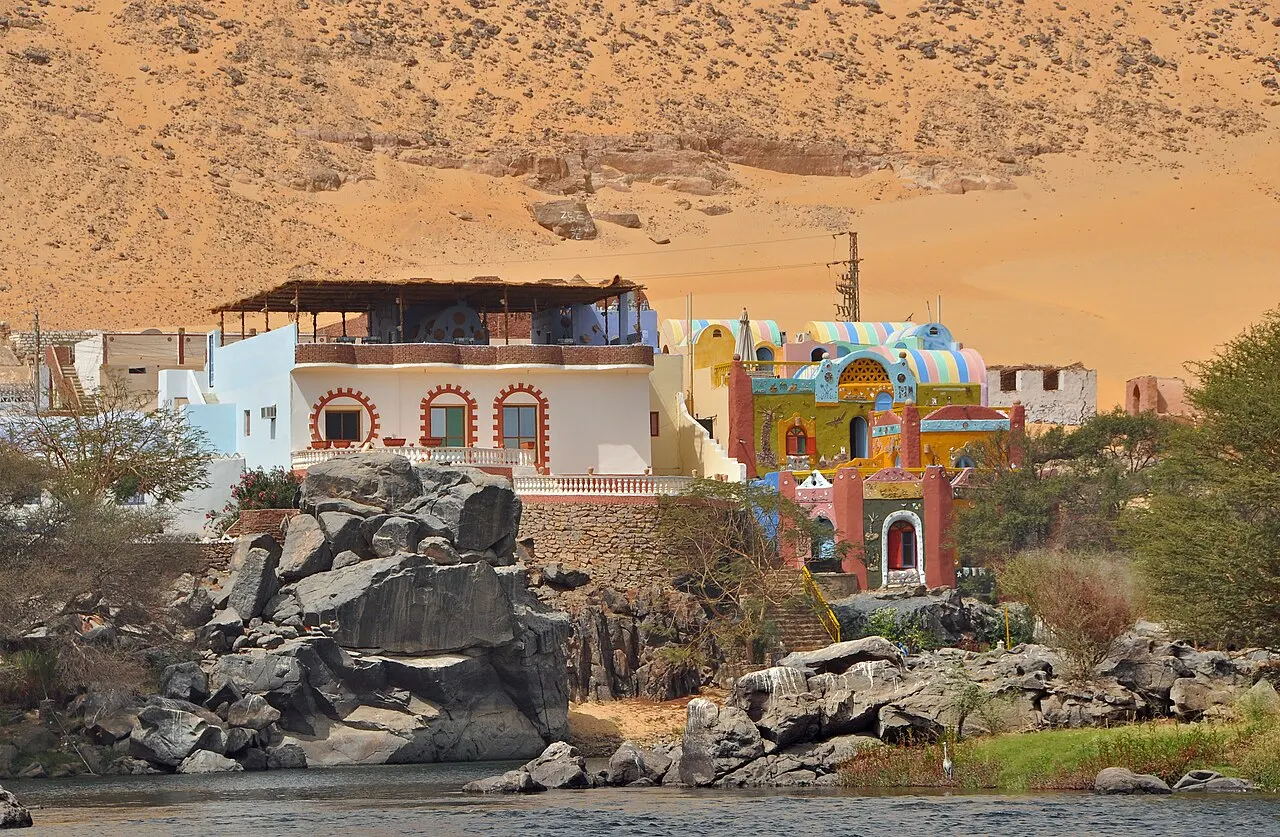 Casas pintadas de blanco, azul, amarillo y rojo, con arcos y adornos de colores, sobre bloques de granito oscuro a orillas del río, con la ladera de arena del desierto detrás.